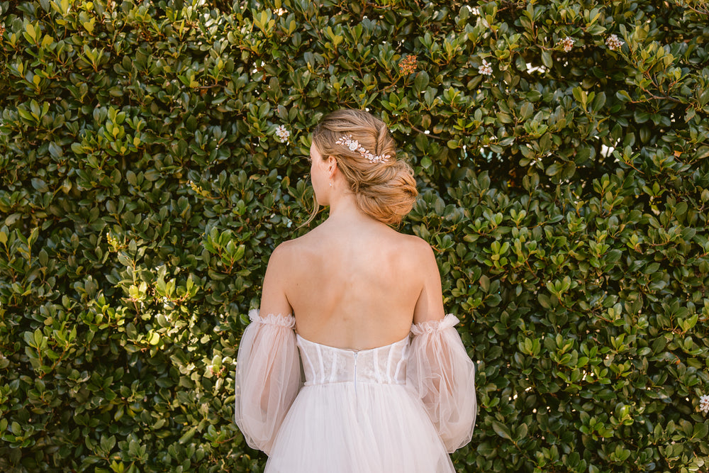 Bride wearing a romantic sleeved blush tulle wedding gown wears a rose gold hair comb with flowers and crystals in a romantic chignon updo